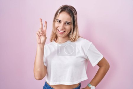 Téléchargez les photos : Jeune femme blonde debout sur fond rose souriant en regardant la caméra montrant des doigts faisant signe de victoire. numéro deux. - en image libre de droit