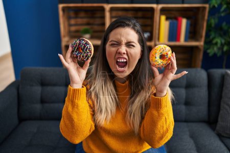 Photo for Young hispanic woman eating doughnuts at home angry and mad screaming frustrated and furious, shouting with anger. rage and aggressive concept. - Royalty Free Image