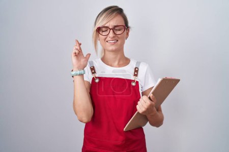 Photo for Young blonde woman wearing waiter uniform holding clipboard gesturing finger crossed smiling with hope and eyes closed. luck and superstitious concept. - Royalty Free Image