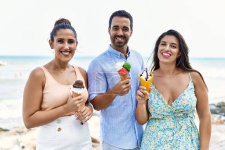 Téléchargez les photos : Trois jeunes amis hispaniques souriant heureux manger de la crème glacée à la plage. - en image libre de droit