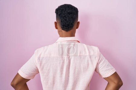 Foto de Joven hombre hispano de pie sobre fondo rosa de pie hacia atrás mirando hacia otro lado con los brazos en el cuerpo - Imagen libre de derechos