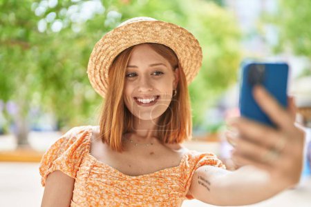 Foto de Young redhead woman tourist wearing summer hat make selfie by smartphone at park - Imagen libre de derechos