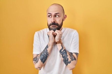 Téléchargez les photos : Jeune homme hispanique avec des tatouages debout sur fond jaune riant nerveux et excité avec les mains sur le menton regardant vers le côté - en image libre de droit