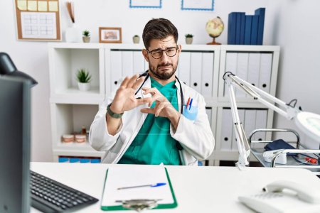 Téléchargez les photos : Young man with beard wearing doctor uniform and stethoscope at the clinic disgusted expression, displeased and fearful doing disgust face because aversion reaction. - en image libre de droit
