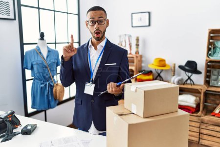Téléchargez les photos : Homme afro-américain travaillant en tant que directeur à la boutique de détail étonné et surpris de regarder vers le haut et pointant du doigt et les bras levés. - en image libre de droit