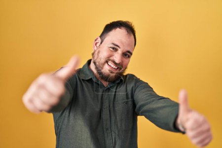 Foto de Plus size hispanic man with beard standing over yellow background approving doing positive gesture with hand, thumbs up smiling and happy for success. winner gesture. - Imagen libre de derechos