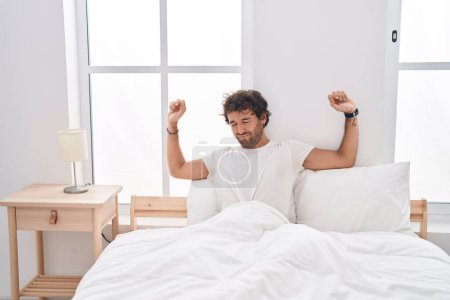 Foto de Young hispanic man waking up stretching arms at bedroom - Imagen libre de derechos