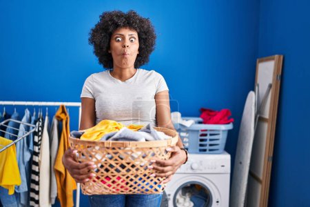 Téléchargez les photos : Black woman with curly hair holding laundry basket making fish face with mouth and squinting eyes, crazy and comical. - en image libre de droit