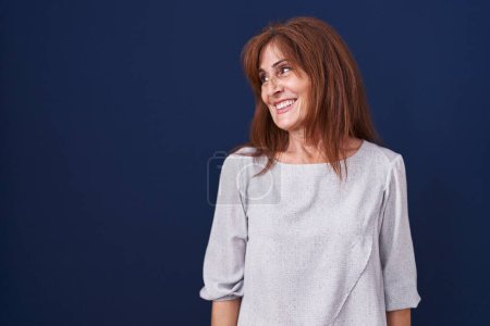 Téléchargez les photos : Middle age woman standing over blue background looking away to side with smile on face, natural expression. laughing confident. - en image libre de droit