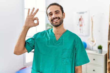 Photo for Young physiotherapist working at pain recovery clinic smiling positive doing ok sign with hand and fingers. successful expression. - Royalty Free Image