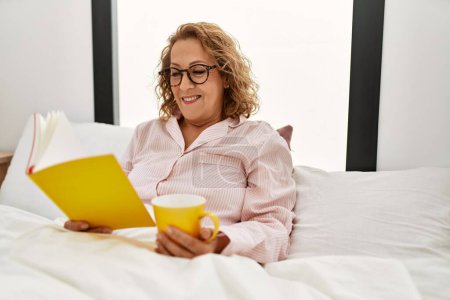 Foto de Mujer caucásica de mediana edad leyendo libro y tomando café sentado en la cama en el dormitorio. - Imagen libre de derechos