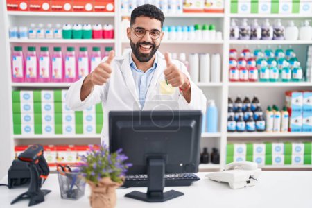 Photo for Hispanic man with beard working at pharmacy drugstore approving doing positive gesture with hand, thumbs up smiling and happy for success. winner gesture. - Royalty Free Image