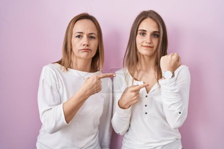 Téléchargez les photos : Middle age mother and young daughter standing over pink background in hurry pointing to watch time, impatience, looking at the camera with relaxed expression - en image libre de droit