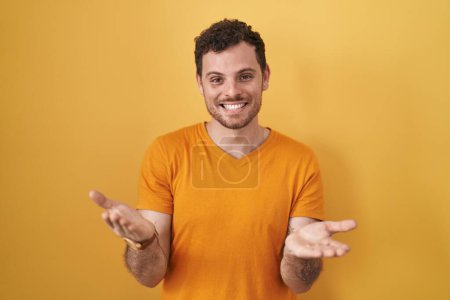 Foto de Joven hombre hispano de pie sobre fondo amarillo sonriente alegre ofreciendo manos dando asistencia y aceptación. - Imagen libre de derechos