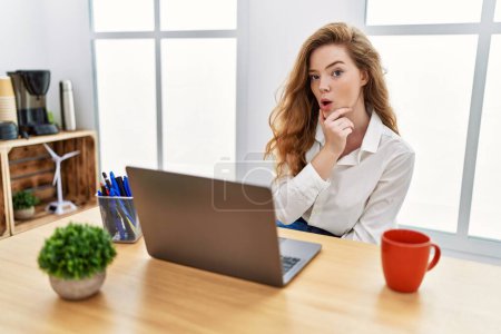 Foto de Mujer joven caucásica que trabaja en la oficina usando computadora portátil que parece fascinada con la incredulidad, sorpresa y expresión asombrada con las manos en la barbilla - Imagen libre de derechos