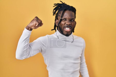 Photo for African man with dreadlocks wearing turtleneck sweater over yellow background angry and mad raising fist frustrated and furious while shouting with anger. rage and aggressive concept. - Royalty Free Image