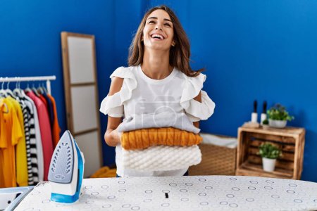 Photo for Young hispanic woman ironing clothes at laundry room holding folded sweaters smiling and laughing hard out loud because funny crazy joke. - Royalty Free Image