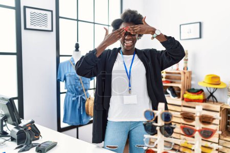 Foto de Mujer joven africana que trabaja como gerente en la boutique minorista cubriendo los ojos con las manos sonriendo alegre y divertido. concepto ciego. - Imagen libre de derechos
