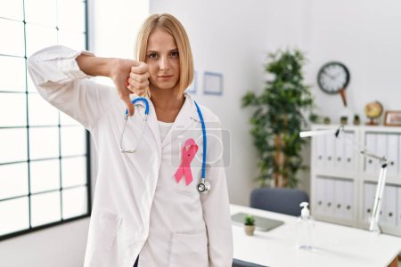 Téléchargez les photos : Young caucasian doctor woman wearing pink cancer ribbon with angry face, negative sign showing dislike with thumbs down, rejection concept - en image libre de droit