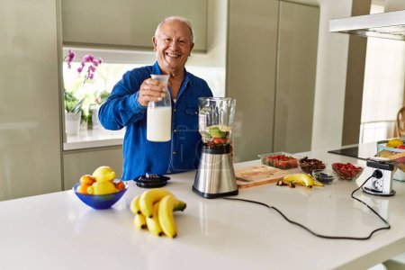 Foto de Hombre mayor sonriendo confiado vertiendo leche en la licuadora en la cocina - Imagen libre de derechos