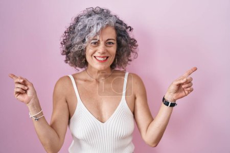 Foto de Mujer de mediana edad con el pelo gris de pie sobre el fondo rosa sonriendo confiado señalando con los dedos a diferentes direcciones. espacio de copia para publicidad - Imagen libre de derechos