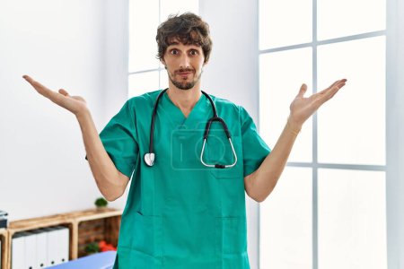 Foto de Young hispanic man wearing doctor uniform and stethoscope at clinic clueless and confused expression with arms and hands raised. doubt concept. - Imagen libre de derechos