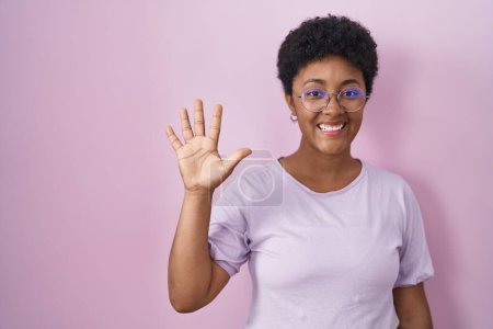 Foto de Joven mujer afroamericana de pie sobre fondo rosa mostrando y apuntando hacia arriba con los dedos número cinco mientras sonríe confiado y feliz. - Imagen libre de derechos