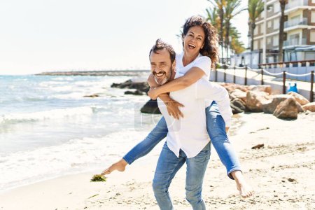 Photo for Middle age hispanic man smiling happy holding woman on his back at the beach. - Royalty Free Image