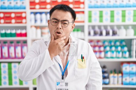 Photo for Chinese young man working at pharmacy drugstore looking fascinated with disbelief, surprise and amazed expression with hands on chin - Royalty Free Image