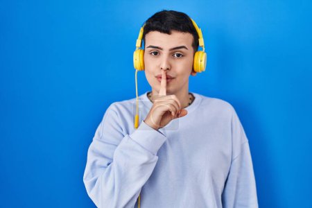 Photo for Non binary person listening to music using headphones asking to be quiet with finger on lips. silence and secret concept. - Royalty Free Image