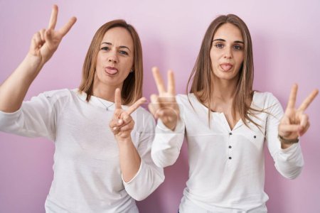 Téléchargez les photos : Middle age mother and young daughter standing over pink background smiling with tongue out showing fingers of both hands doing victory sign. number two. - en image libre de droit