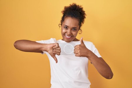 Photo for Young hispanic woman with curly hair standing over yellow background doing thumbs up and down, disagreement and agreement expression. crazy conflict - Royalty Free Image