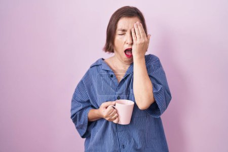 Foto de Mujer hispana de mediana edad bebiendo una taza de café bostezando cansada cubriendo media cara, ojos y boca con la mano. cara duele en el dolor. - Imagen libre de derechos