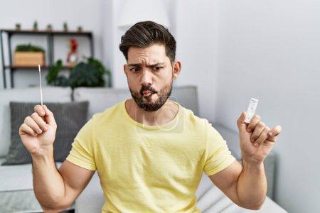 Photo for Young man with beard holding coronavirus infection nasal test making fish face with mouth and squinting eyes, crazy and comical. - Royalty Free Image