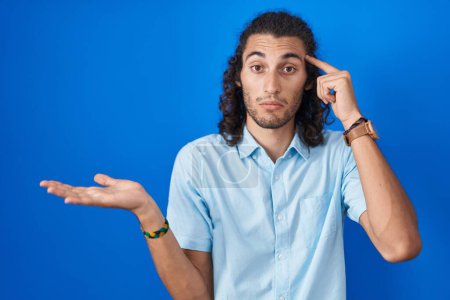 Téléchargez les photos : Young hispanic man standing over blue background confused and annoyed with open palm showing copy space and pointing finger to forehead. think about it. - en image libre de droit