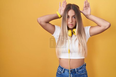 Foto de Young blonde woman standing over yellow background wearing headphones doing bunny ears gesture with hands palms looking cynical and skeptical. easter rabbit concept. - Imagen libre de derechos