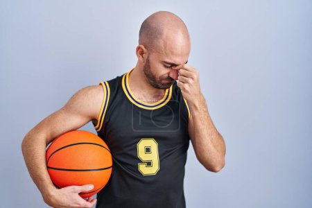 Young bald man with beard wearing basketball uniform holding ball tired rubbing nose and eyes feeling fatigue and headache. stress and frustration concept. 