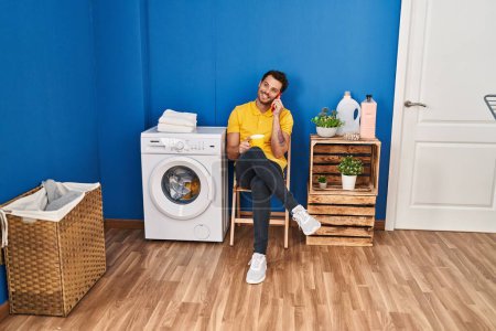 Téléchargez les photos : Jeune homme hispanique boire du café parler sur le smartphone wating pour machine à laver à la buanderie - en image libre de droit