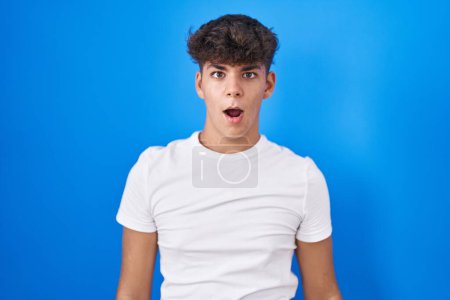 Foto de Adolescente hispano de pie sobre fondo azul en la cara de choque, con aspecto escéptico y sarcástico, sorprendido con la boca abierta - Imagen libre de derechos