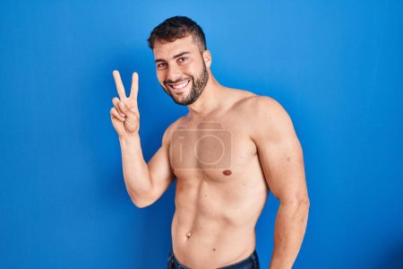 Téléchargez les photos : Bel homme hispanique debout torse nu souriant regardant la caméra montrant les doigts faisant signe de victoire. numéro deux. - en image libre de droit