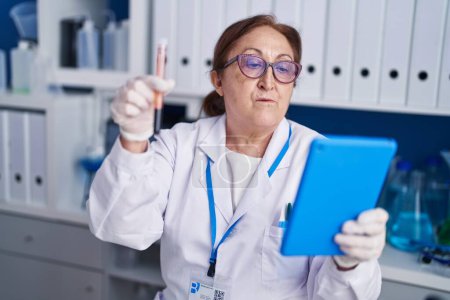 Photo for Senior woman scientist using touchpad holding blood test tube at laboratory - Royalty Free Image