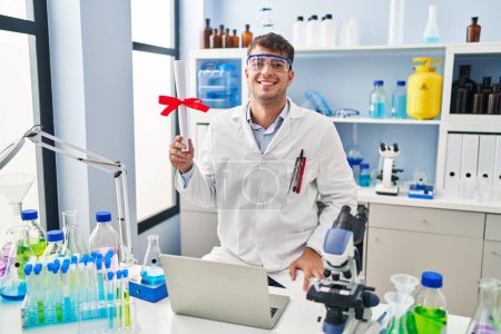 Téléchargez les photos : Jeune homme hispanique travaillant au laboratoire scientifique titulaire d'un diplôme regardant positif et heureux debout et souriant avec un sourire confiant montrant les dents - en image libre de droit