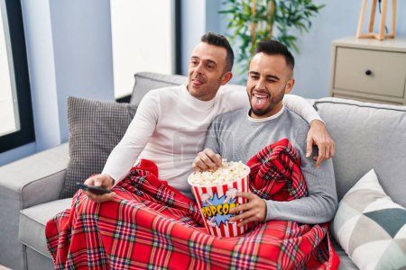 Foto de Pareja homosexual comiendo palomitas de maíz viendo televisión sacando la lengua feliz con expresión divertida. - Imagen libre de derechos