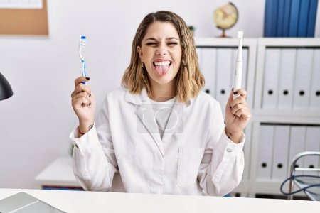 Junge hispanische Zahnärztin hält elektrische Zahnbürste und Zahnbürste in der Klinik und streckt mit lustigem Gesichtsausdruck die Zunge heraus. 