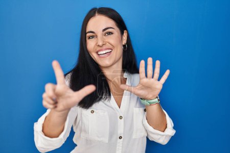 Foto de Mujer hispana joven de pie sobre fondo azul mostrando y señalando hacia arriba con los dedos número siete mientras sonríe confiado y feliz. - Imagen libre de derechos