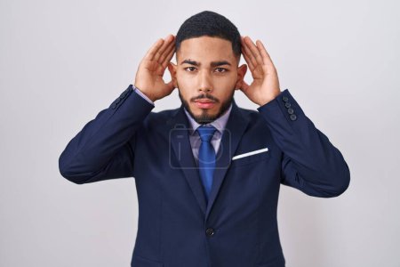 Téléchargez les photos : Jeune homme hispanique portant un costume d'affaires et cravate en essayant d'entendre les deux mains sur le geste de l'oreille, curieux de commérages. problème auditif, sourd - en image libre de droit
