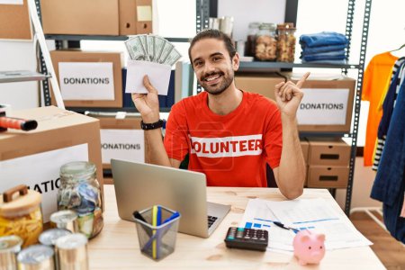 Photo for Young hispanic man holding money at donations stand smiling happy pointing with hand and finger to the side - Royalty Free Image