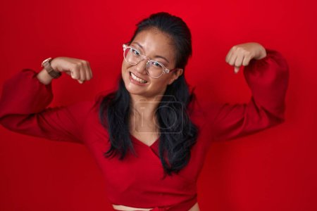 Foto de Asian young woman standing over red background showing arms muscles smiling proud. fitness concept. - Imagen libre de derechos