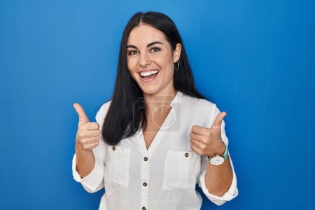 Photo for Young hispanic woman standing over blue background success sign doing positive gesture with hand, thumbs up smiling and happy. cheerful expression and winner gesture. - Royalty Free Image