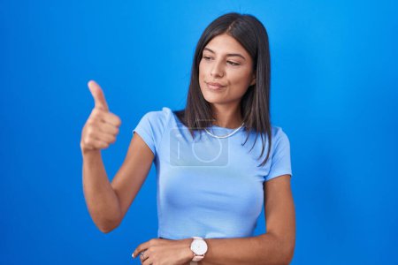 Téléchargez les photos : Brunette young woman standing over blue background looking proud, smiling doing thumbs up gesture to the side - en image libre de droit
